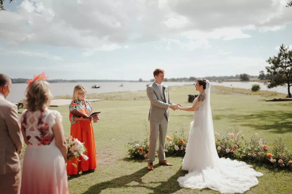Wedding celebrant Eleanor Willock stands in the ceremony space by the water's edge, a bride and groom holding hands in front of her