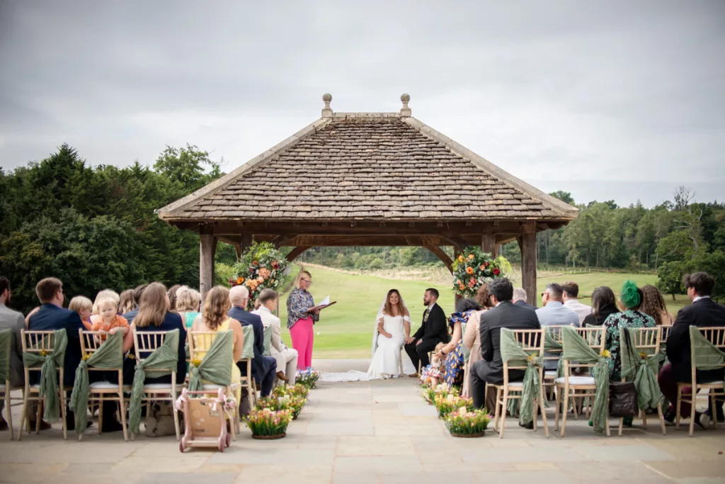 A bride and groom are seated under the pagoda at Bowood House, Eleanor the celebrant is speaking to them from the left hand side whilst the guests sit in rows watching 